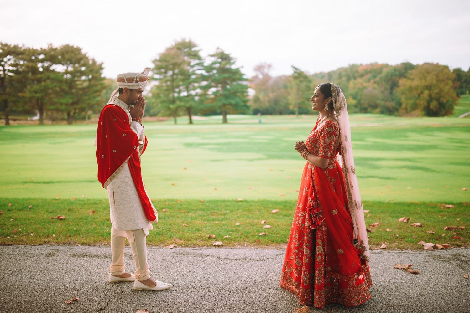A couple in traditional South Asian wedding attire stand together on a path overlooking a green golf course in autumn.