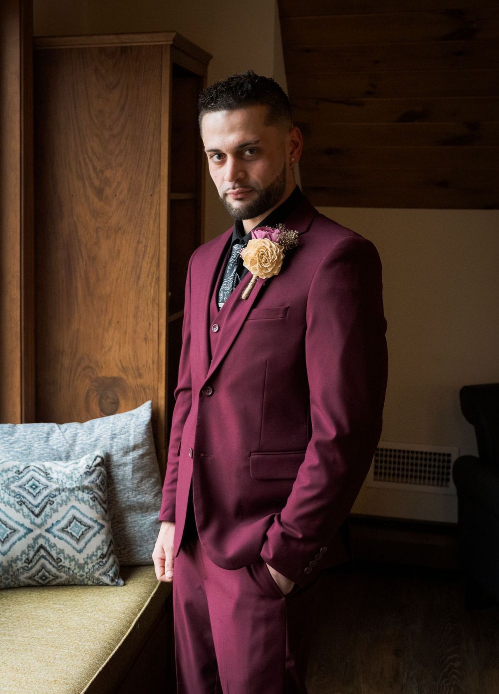 Groom in burgundy three-piece suit with floral boutonniere posing elegantly before wedding ceremony.