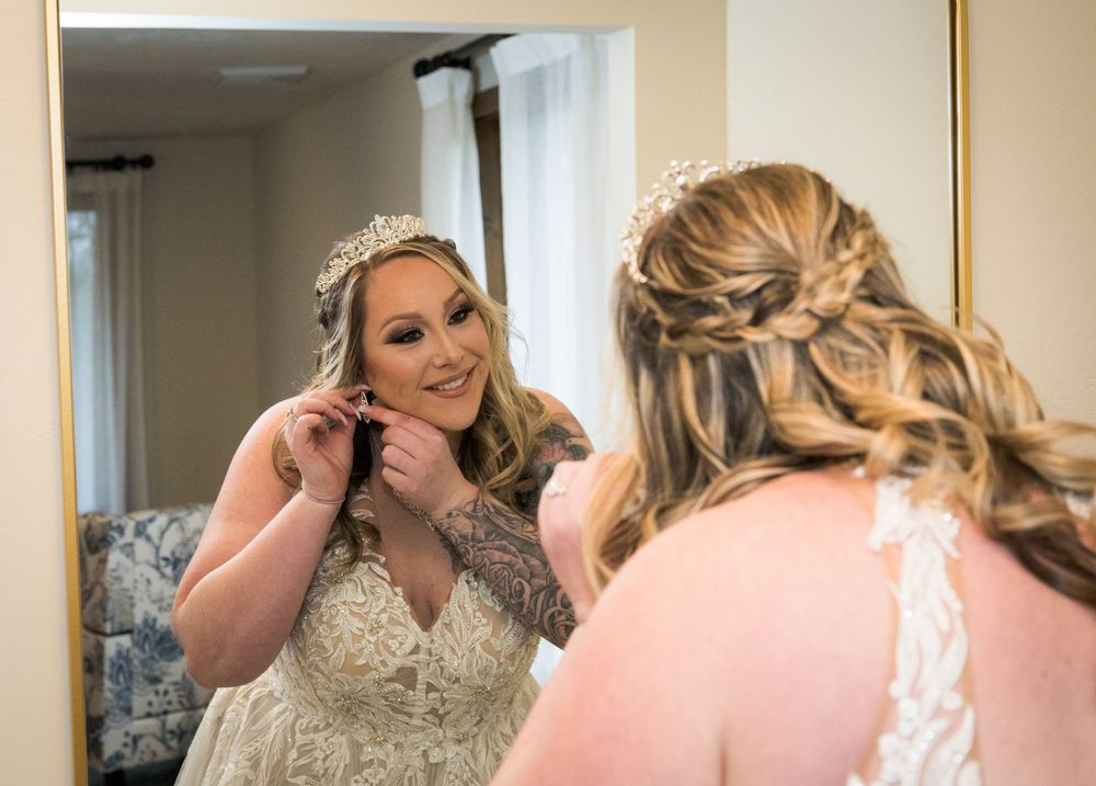 Bride in lace wedding gown adjusting earrings while admiring reflection in mirror before ceremony.