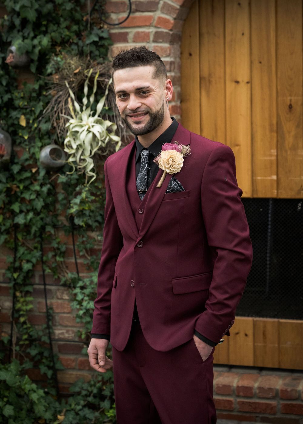 Man in burgundy three-piece suit with floral boutonniere posing outdoors near brick wall and wooden door.