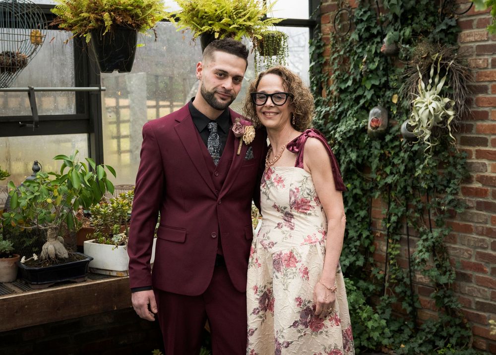 Couple posing together at a garden wedding, man in burgundy suit and woman in floral dress wearing glasses.
