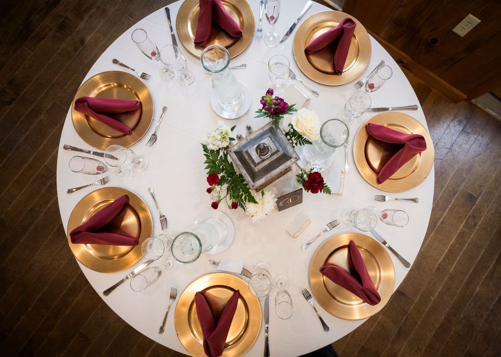Elegant round wedding reception table set with gold charger plates, burgundy napkins, crystal glassware, and a floral centerpiece.