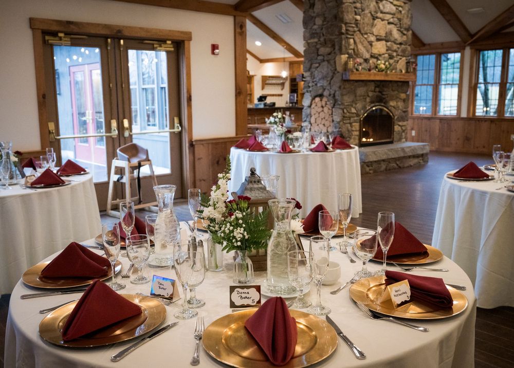 Elegant wedding reception room with round tables set with gold chargers, burgundy napkins, and floral centerpieces near a stone fireplace.