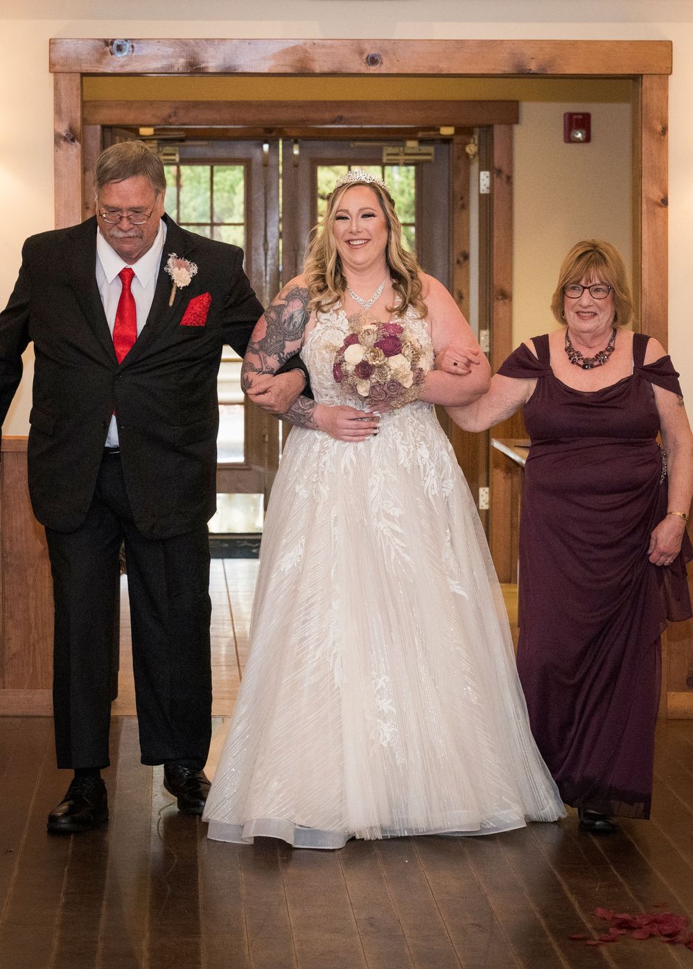 Bride in lace gown walks down aisle escorted by parents at rustic wedding venue.