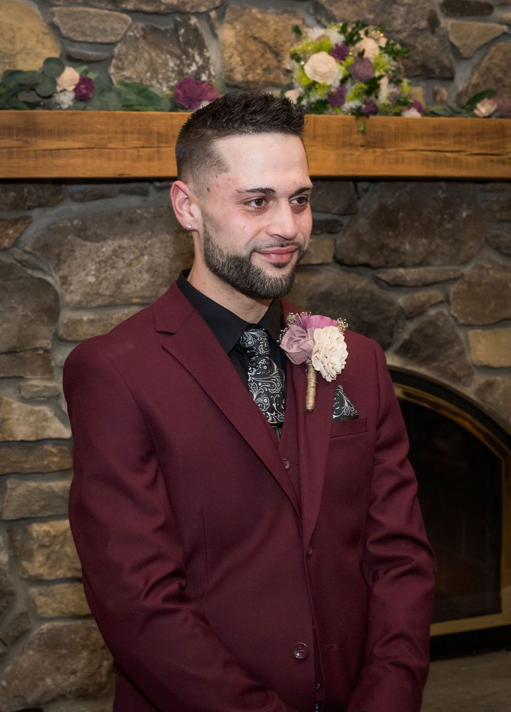 Groom in burgundy suit with boutonniere stands before stone fireplace at wedding ceremony.