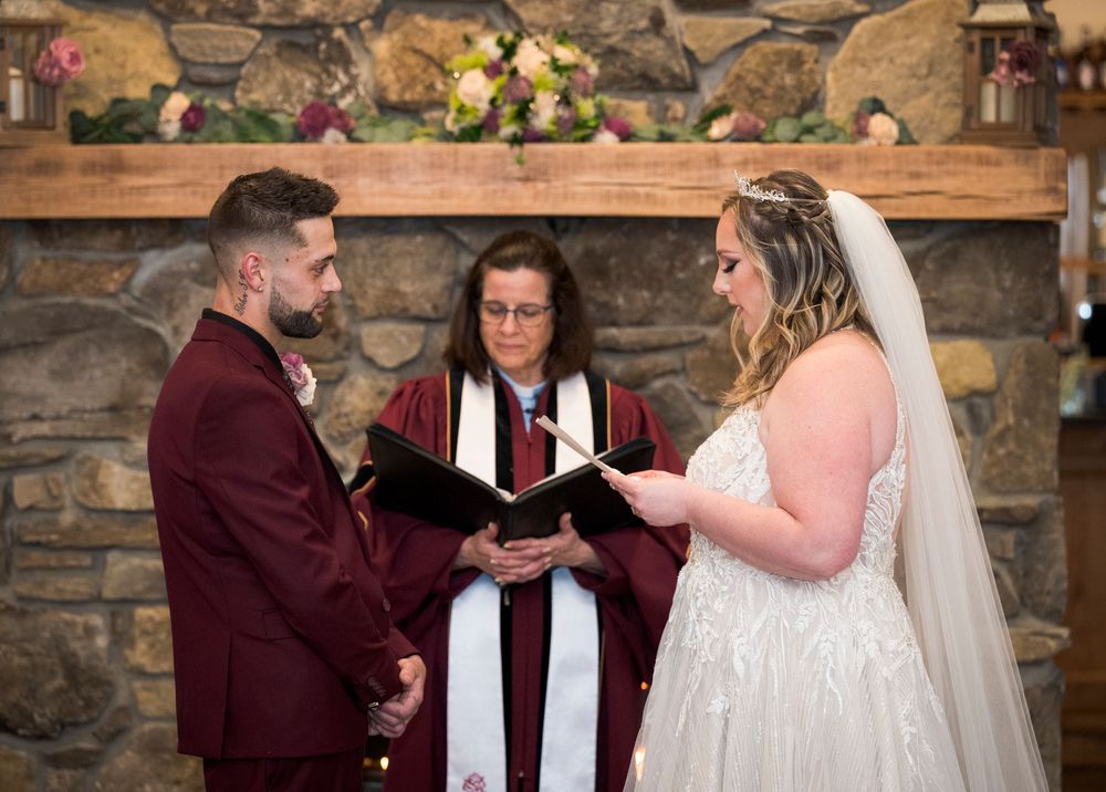 Bride and groom exchange vows before a female officiant at a rustic stone fireplace wedding ceremony.