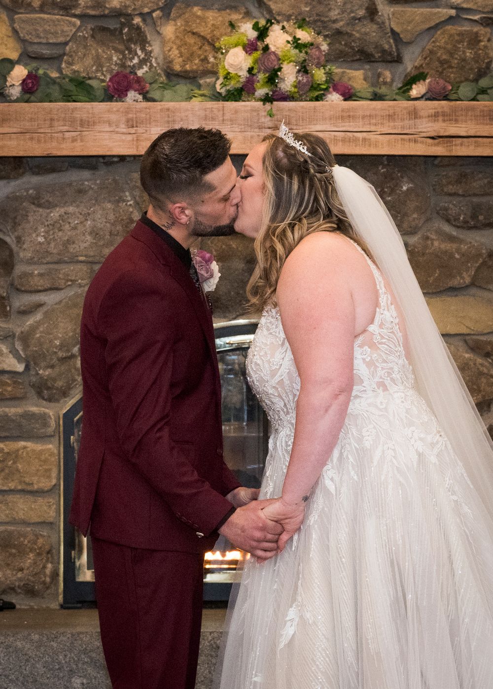 Bride and groom share first kiss at stone fireplace altar, groom in burgundy suit, bride in lace gown with veil.