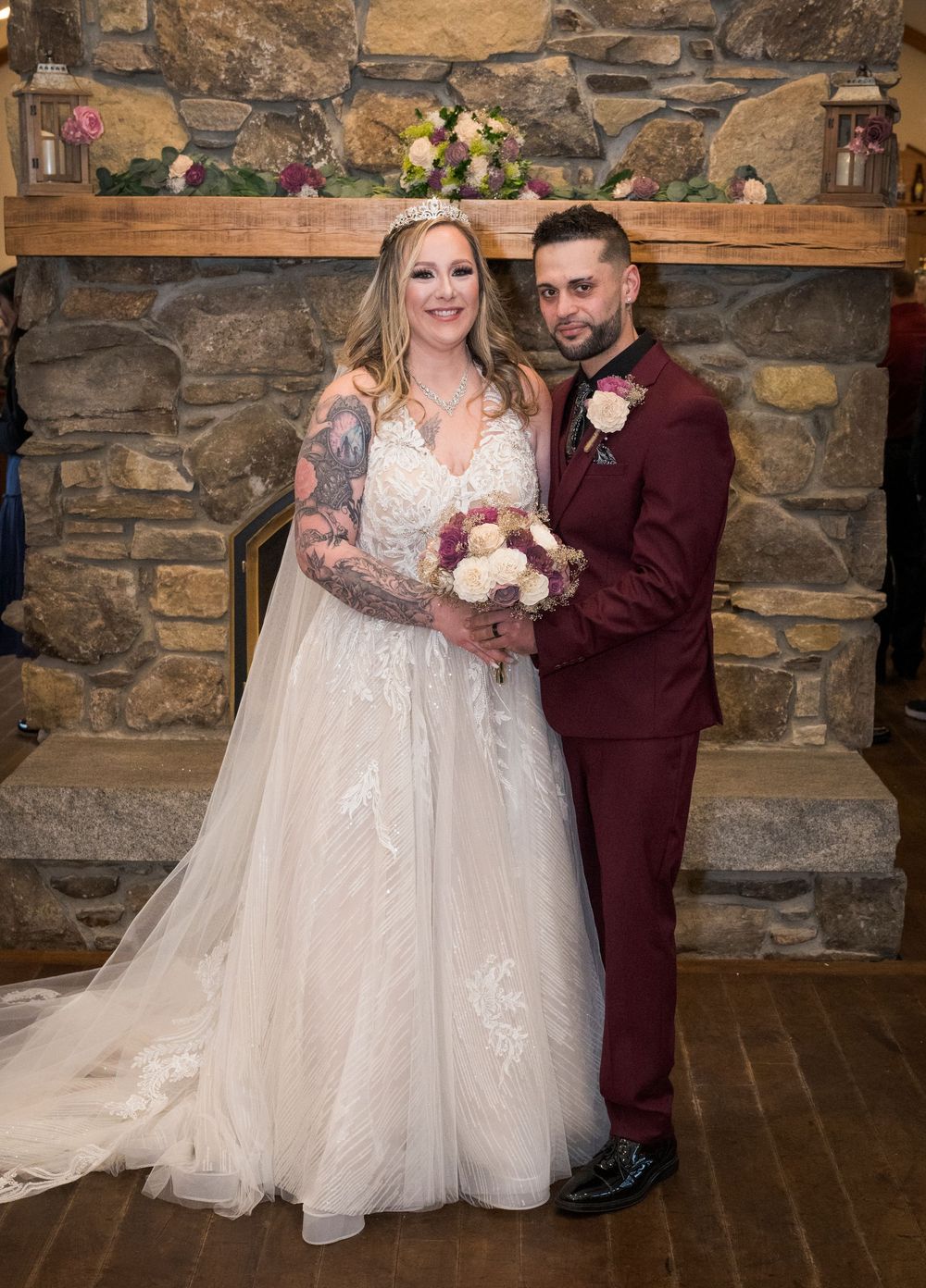 Bride in white lace gown and groom in burgundy suit pose together in front of a stone fireplace on their wedding day.