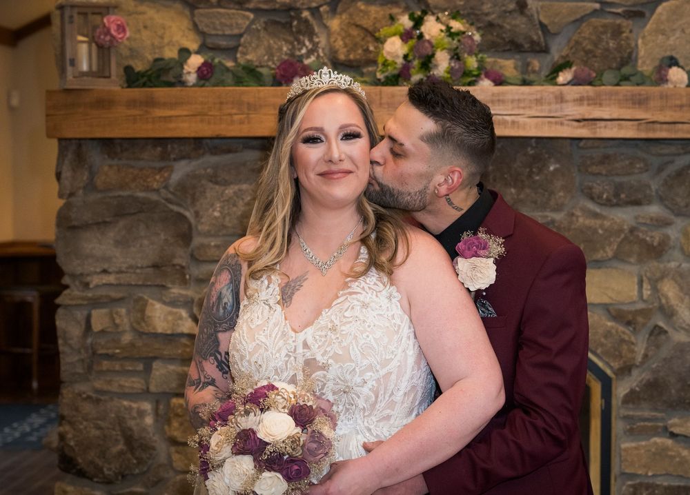 Bride in white lace gown with tiara holds bouquet as groom in burgundy suit kisses her cheek by stone fireplace.