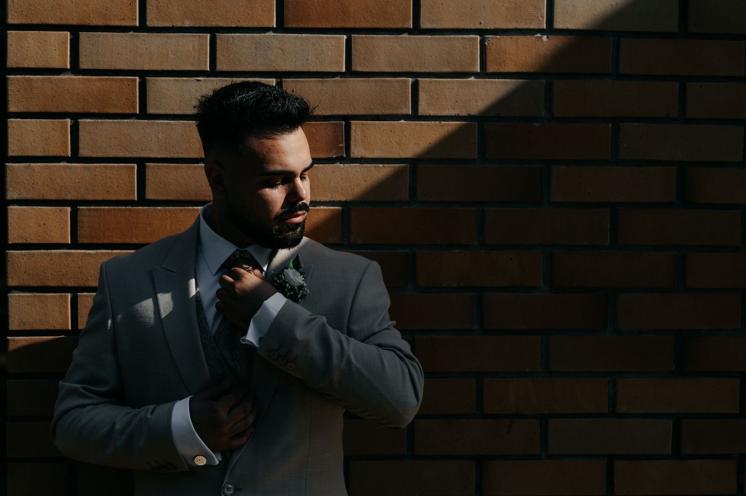 A person in a creamy suit jacket leans against a brick wall while looking thoughtfully to the side.