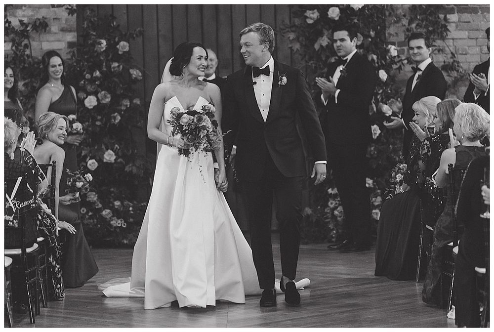 Wedding party walks down aisle during ceremony while guests look on in formal indoor venue.