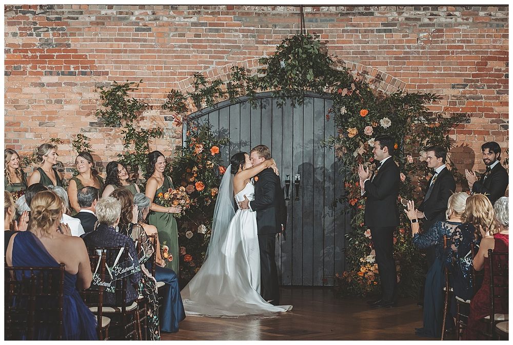 Newlyweds share first kiss against brick wall with floral arch and guests watching during indoor ceremony.