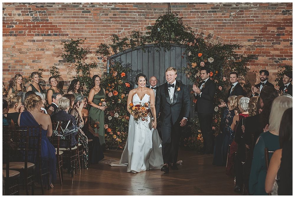 A wedding ceremony in a rustic brick venue with guests seated on either side of the aisle and greenery on the wall.