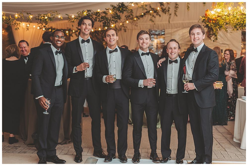 Groomsmen in black tuxedos and bowties pose together at an elegant wedding reception with string lights in background.