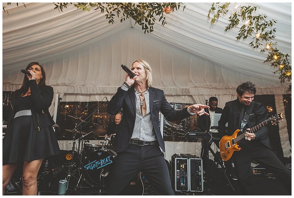 Live band performs under white tent with string lights and autumn decor at evening wedding reception.