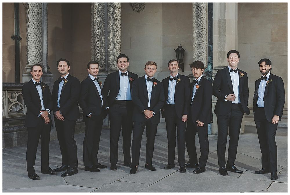 A group of nine groomsmen in black suits and bow ties pose together against an ornate stone building exterior.