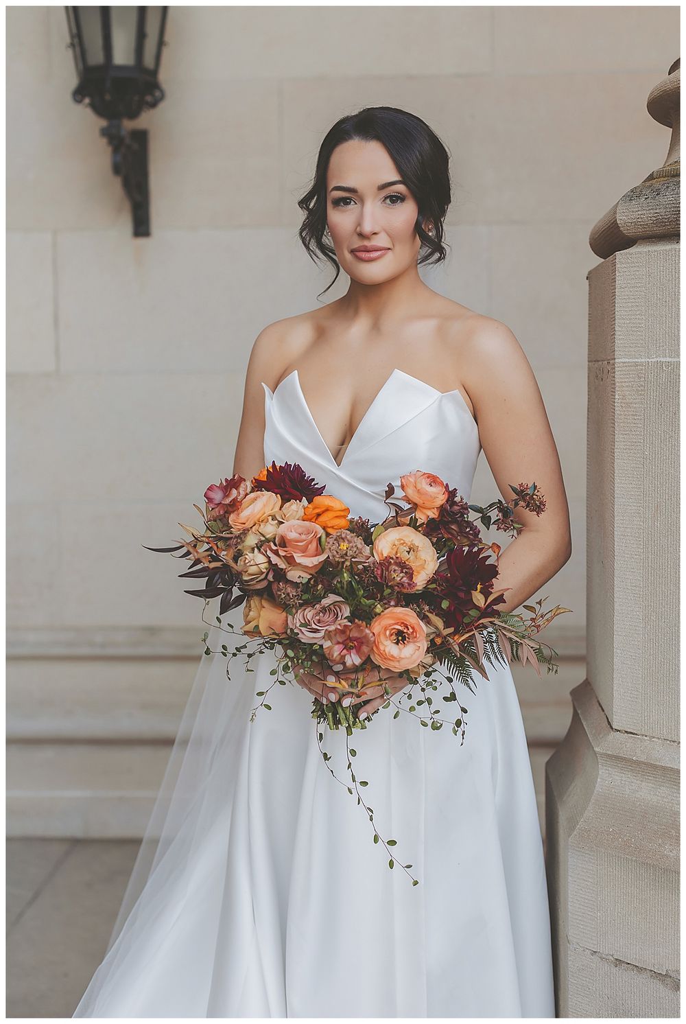 A bride in a white dress holds a vibrant autumn-colored bouquet featuring peach and orange flowers against a stone wall.