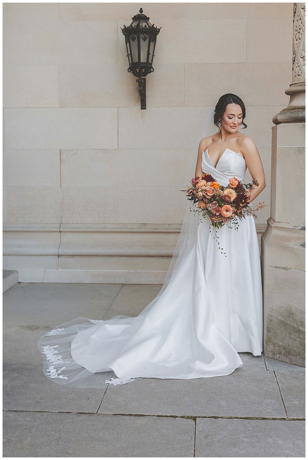 A bride in a white strapless wedding gown holds an autumn-colored bouquet while posing against a stone wall.