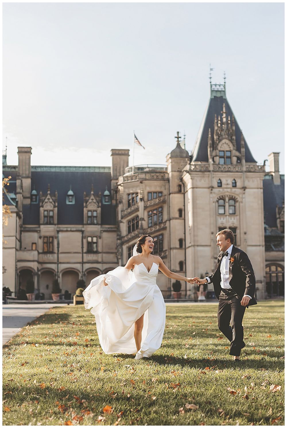 A couple in wedding attire runs playfully on the lawn in front of the grand Biltmore Estate mansion in Asheville.