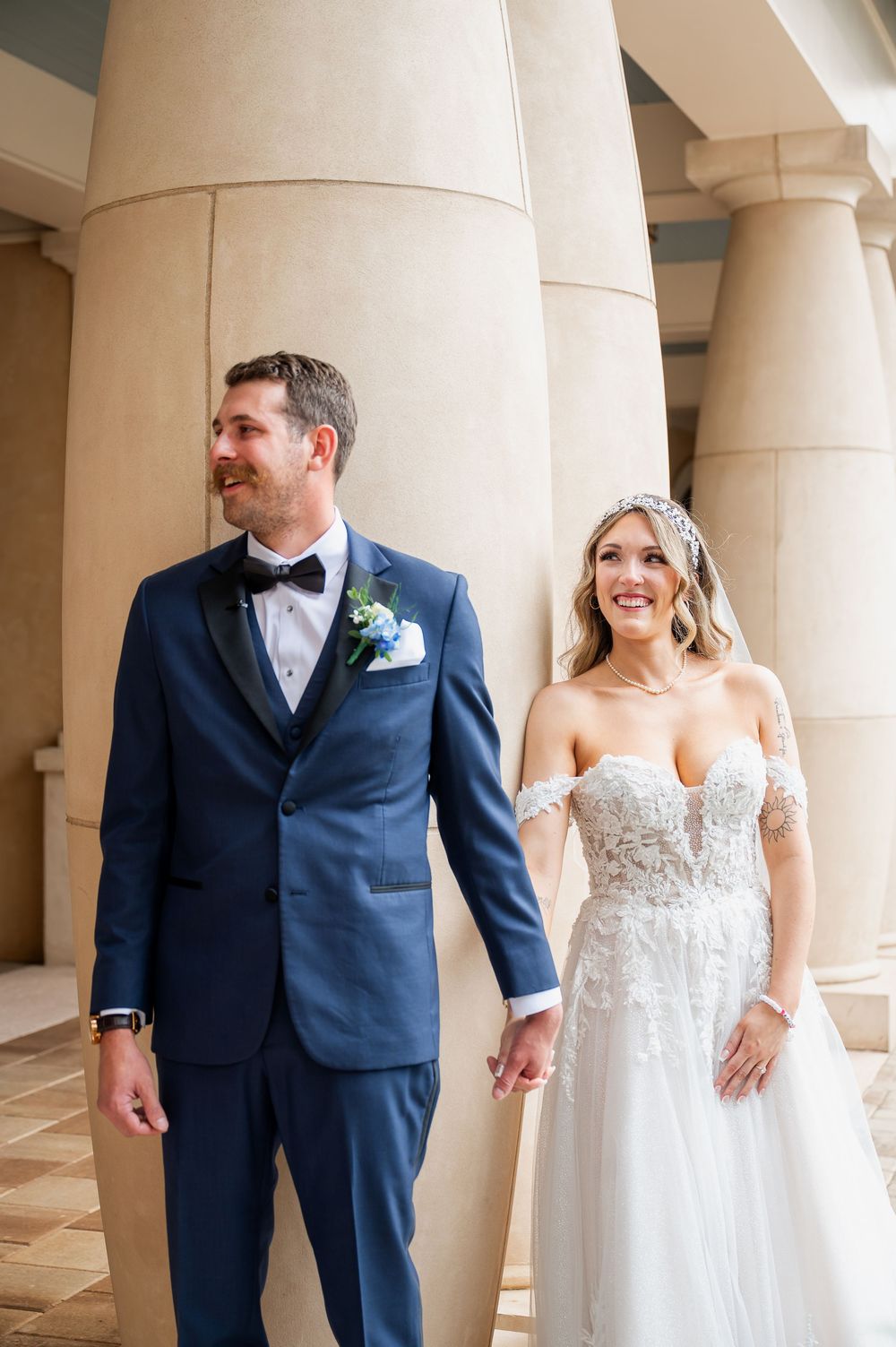 Happy couple in formal wedding attire walk together among architectural columns.