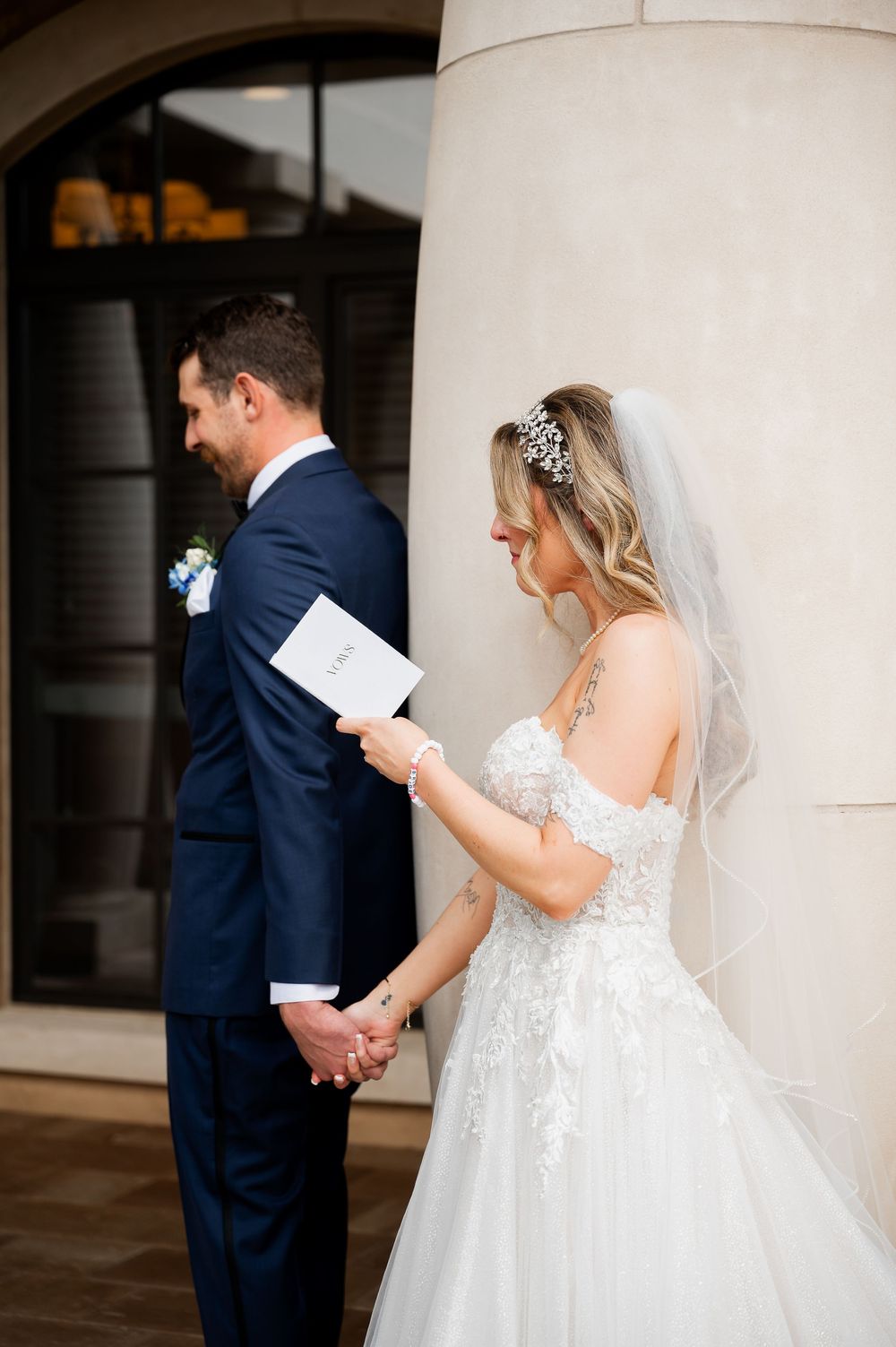 Bride in lace gown reads letter while groom waits during first look sequence.
