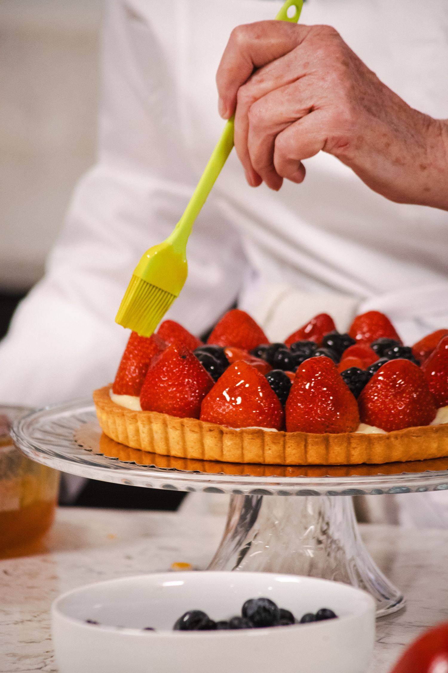 Chef brushing pastry glaze onto fresh fruit tart with strawberries and blueberries on glass cake stand.