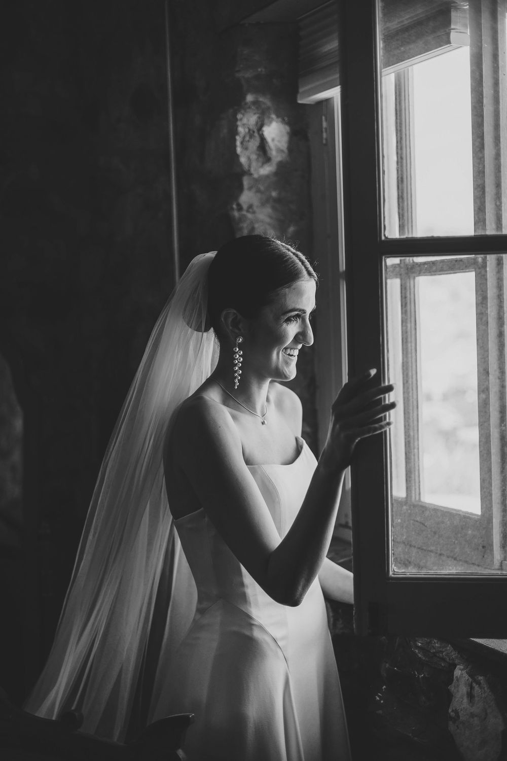 Black and white photo series of a bride in a flowing veil looking through windows in a rustic interior setting.