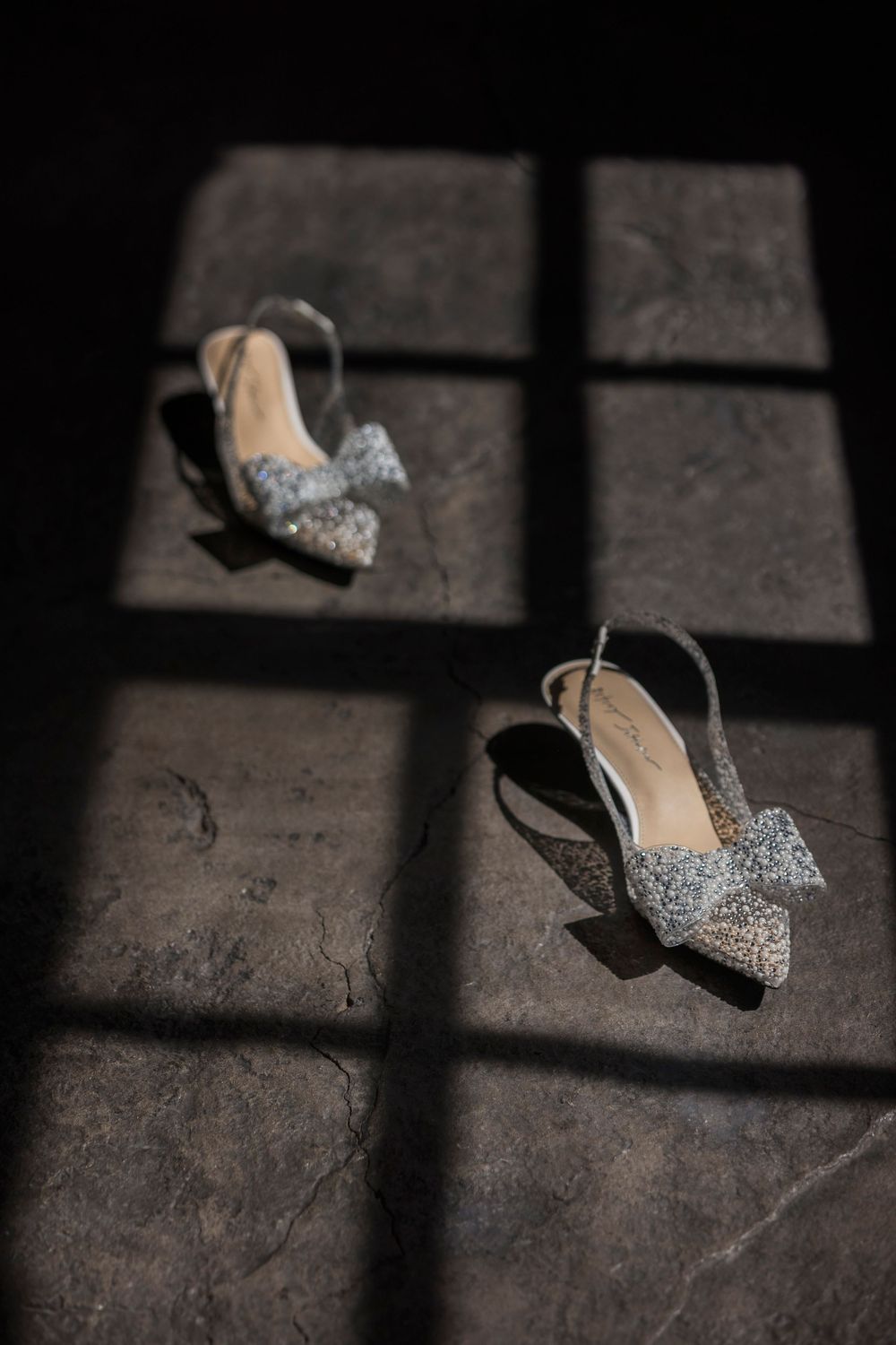 A sparkly silver high heel bridal shoe casts dramatic shadows on a textured concrete floor in dramatic lighting.