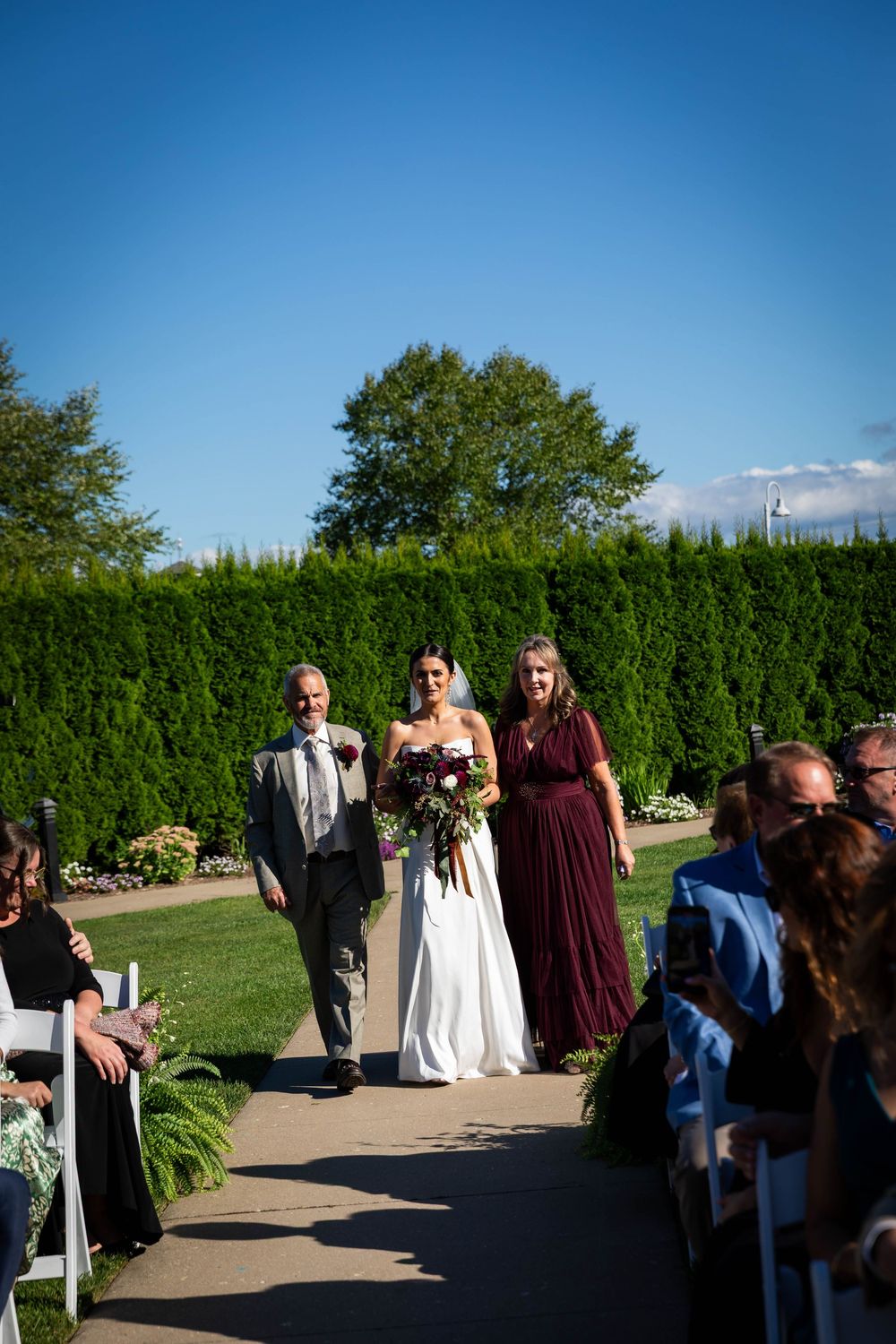 Mother and father of the bride walking her down the aisle outdoors in the summer sun