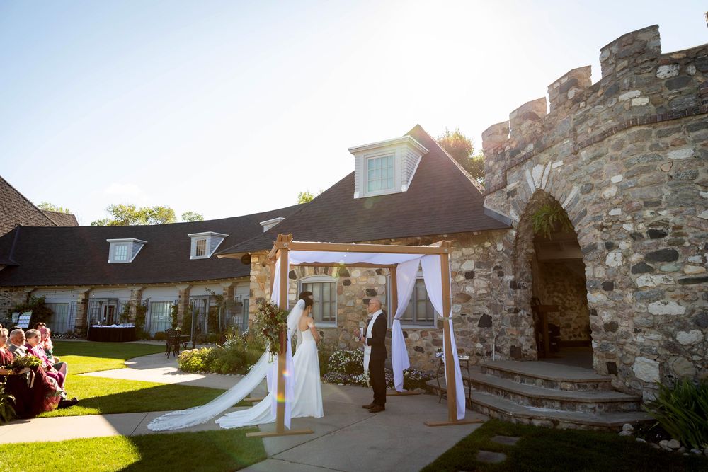A wedding couple stands in front of a rustic stone wall as sunlight streams through trees during their outdoor ceremony.