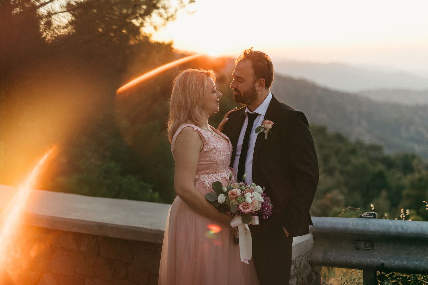A romantic sunset moment captured on a mountain overlook with a pink dress and bouquet of flowers.