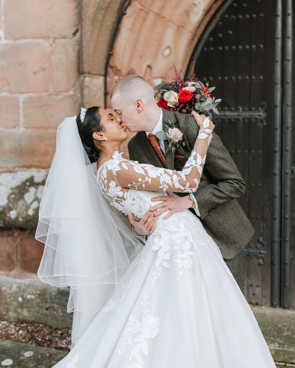 A romantic wedding couple shares a tender kiss outdoors in front of a stone church wall.