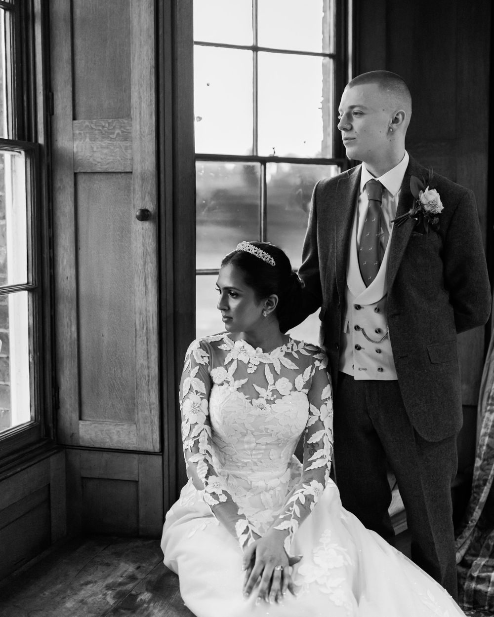 Black and white wedding photo showing a bride in long sleeve lace dress seated by a window in elegant interior setting.