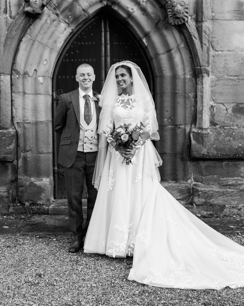 A vintage black and white wedding photograph showing a bride and companion standing in front of a gothic stone archway.