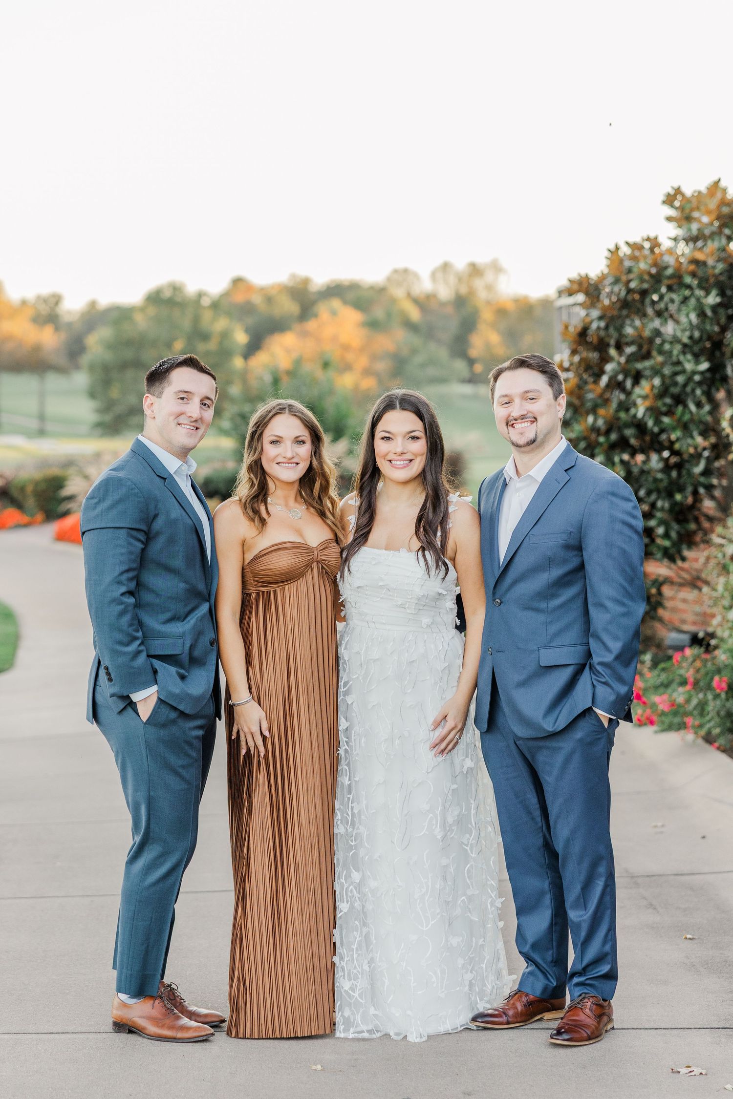 A group poses outdoors in formal attire with navy suits and brown and white dresses against an autumn backdrop.