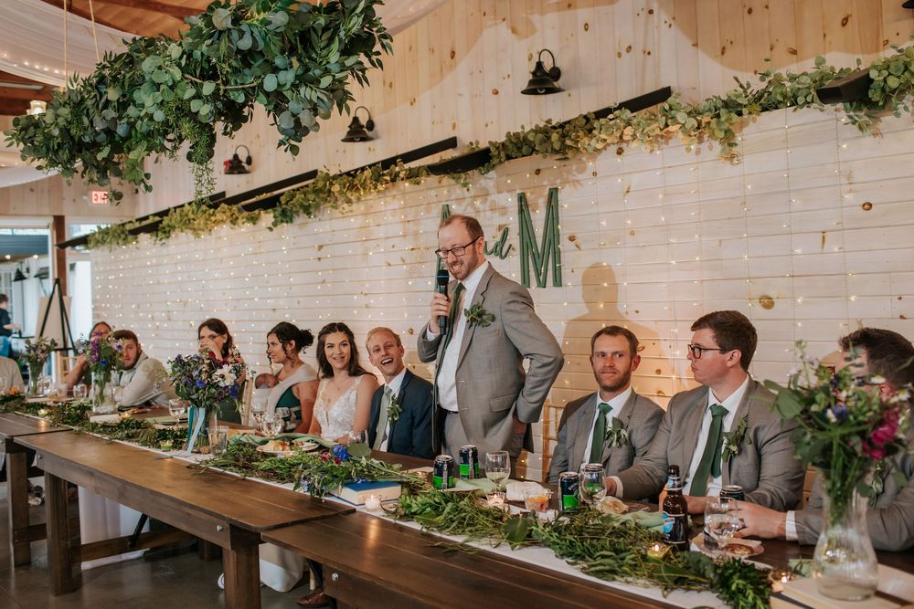 Wedding party seated at long rustic table decorated with greenery under wooden beams.