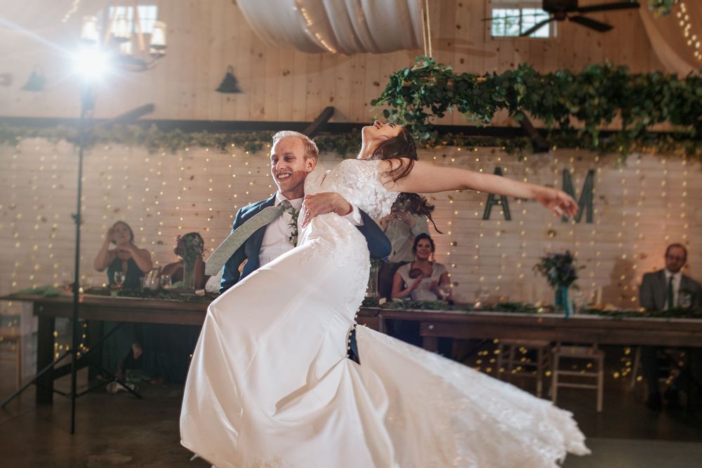 Wedding couple dances together in a rustic venue decorated with twinkling lights and greenery.