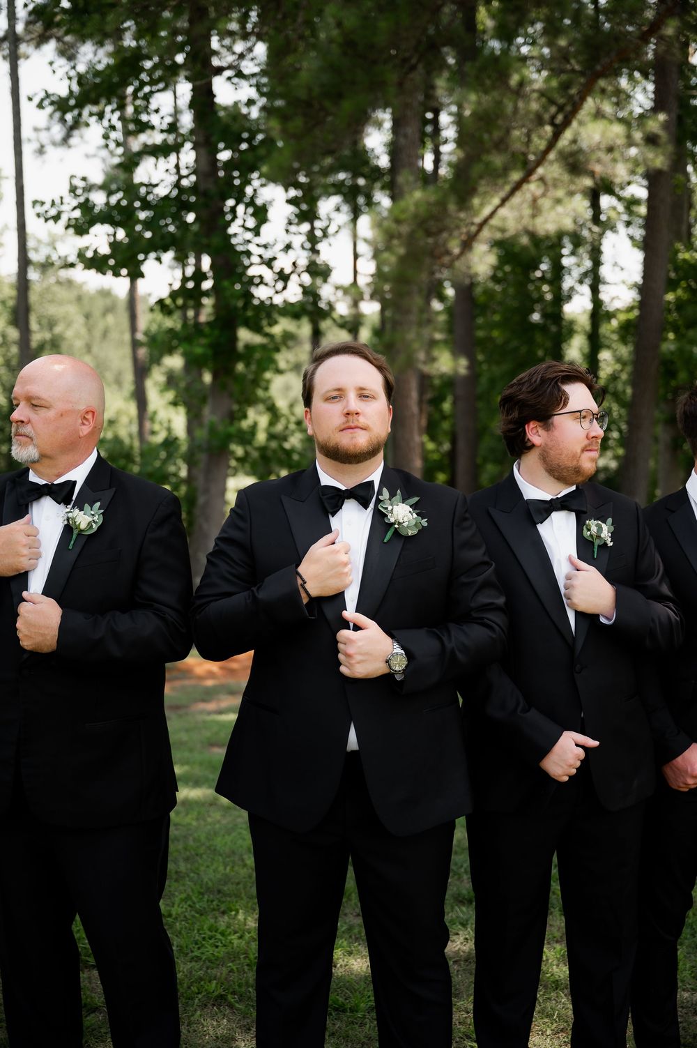 Groomsmen in black tuxedos adjust their bowties while posing in a wooded setting.