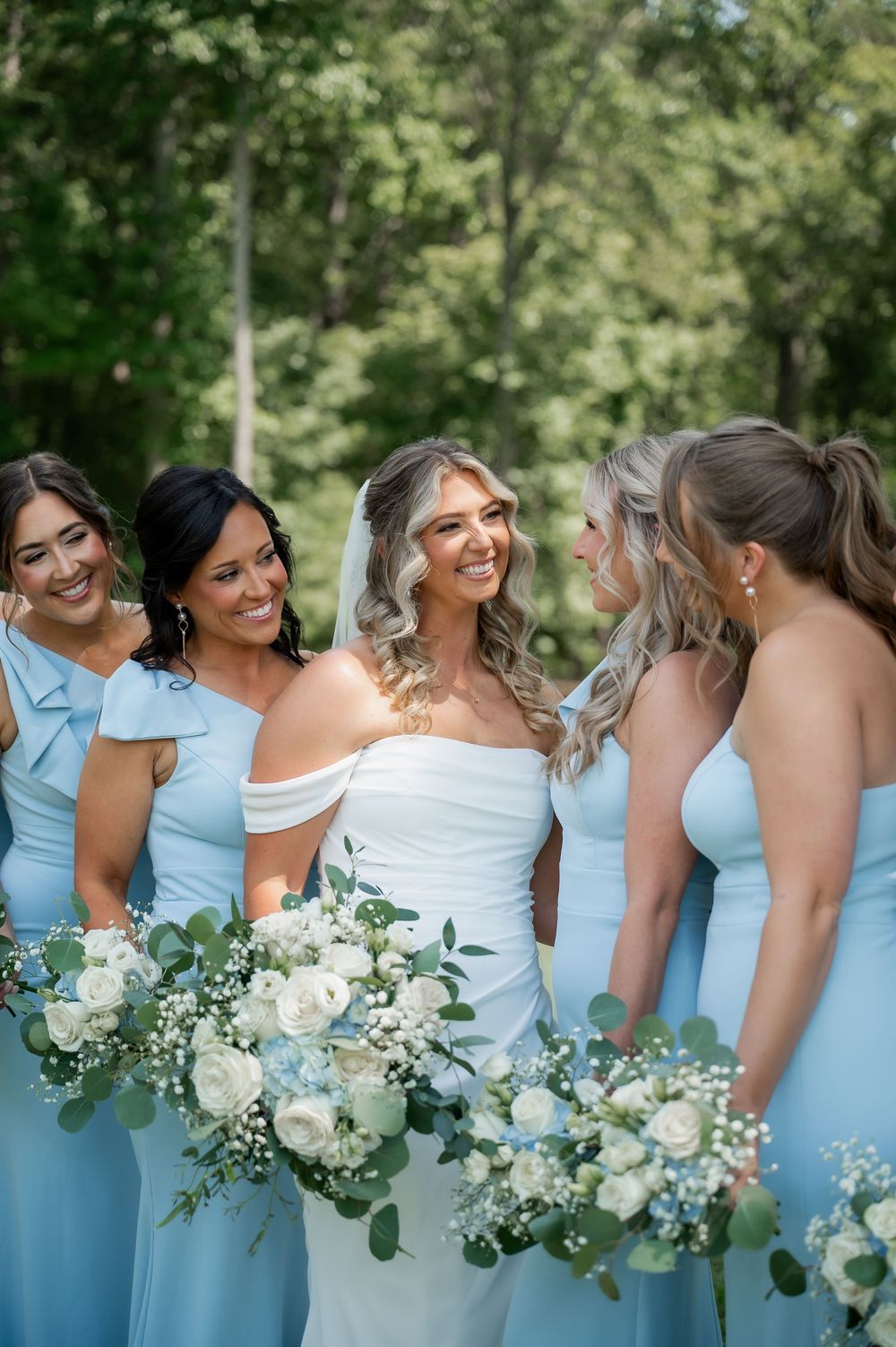 Bridesmaids in light blue dresses hold white floral bouquets while sharing a candid moment.