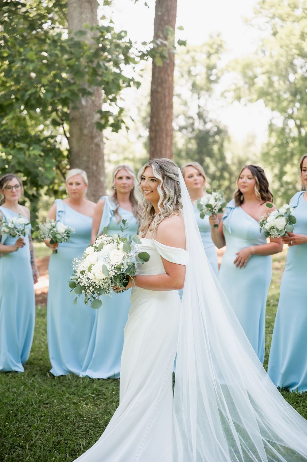 Bride in white gown poses with bridesmaids wearing flowing light blue dresses in an outdoor setting.