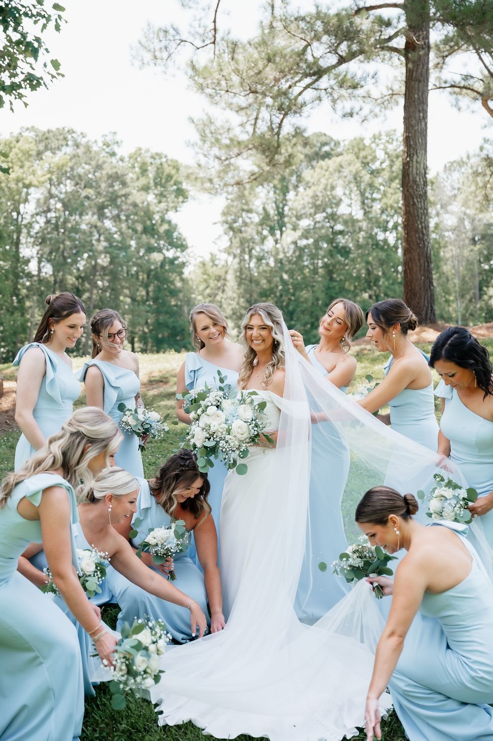 Bridesmaids in light blue dresses help arrange the bride's wedding gown train outdoors.