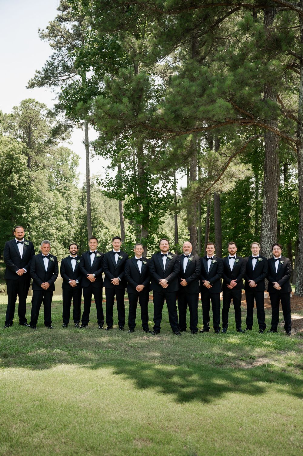 Groomsmen in black tuxedos stand in a line against a backdrop of tall pine trees.