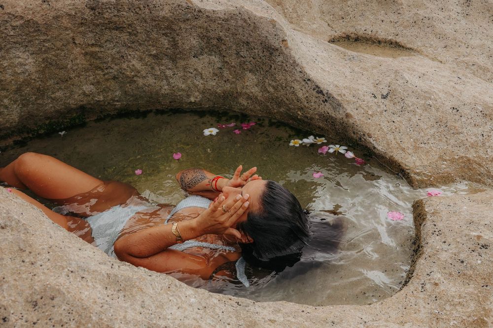 Woman lying in a natural rock pool with flower petals floating on water at a beach.