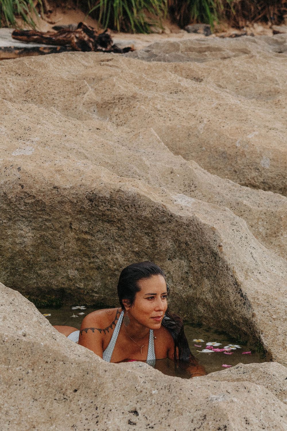 Woman in white bikini relaxing in a natural rock pool carved into sandy stone at beach.