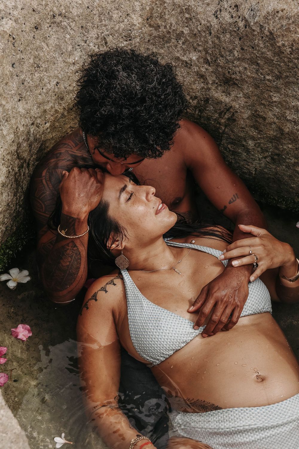 Couple sharing an intimate moment in a natural rock pool with flower petals floating in water.
