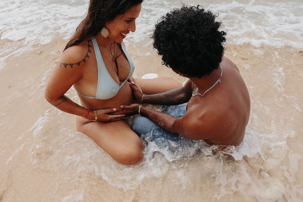 Couple sitting together in ocean waves on a tropical beach, smiling and holding hands.