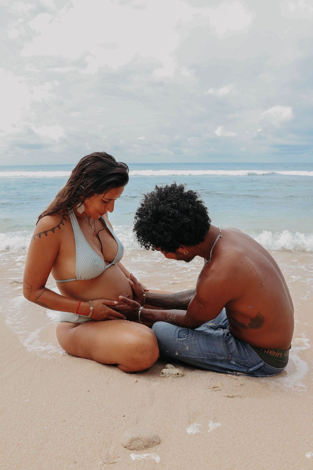 Pregnant woman in white bikini with partner touching baby bump while sitting on sandy beach.