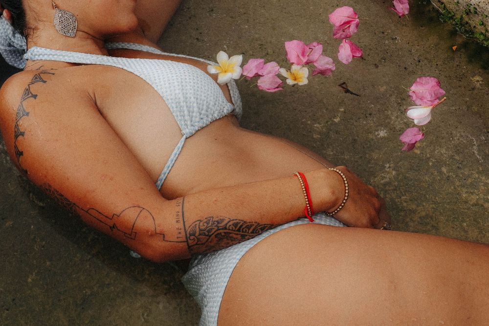 Close-up of tattooed woman in white bikini lying near pink flower petals with red anklet.