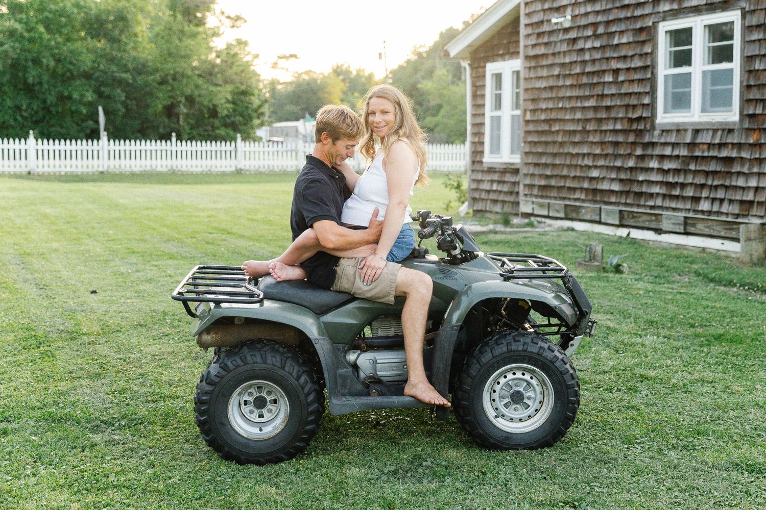 A couple sits together on a parked ATV on a grassy lawn next to a rustic shingle-sided house.