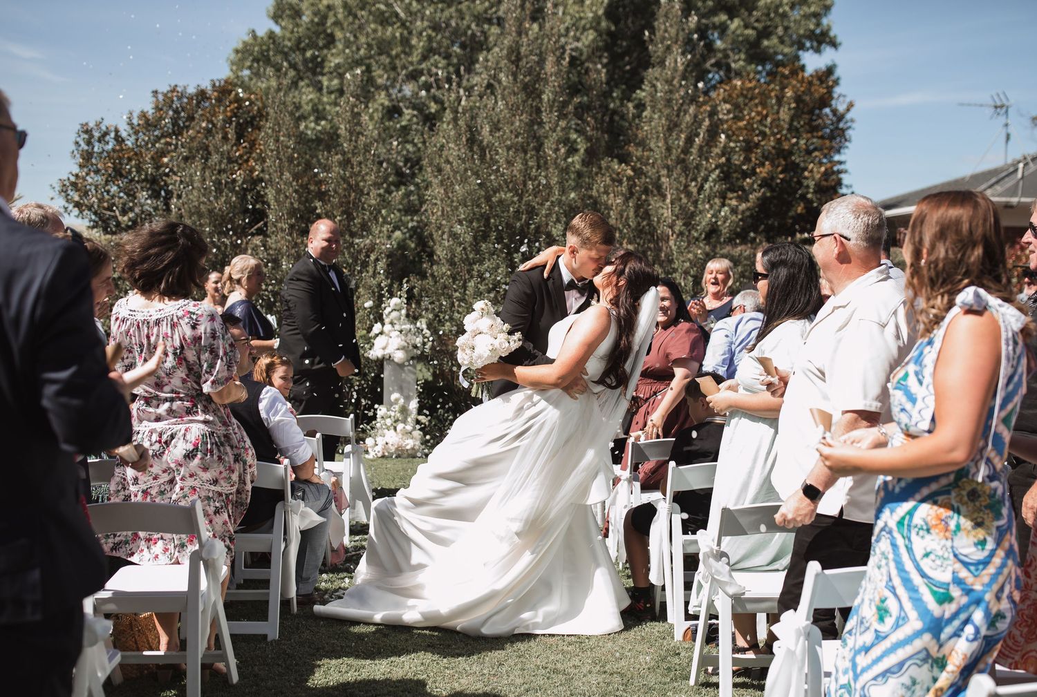 Wedding guests celebrate during an outdoor reception with white chairs and tables under trees on a sunny day.